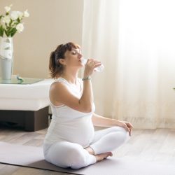 Una mujer embarazada está sentada con las piernas cruzadas sobre una colchoneta de yoga, bebiendo agua hidrogenada en una habitación luminosa llena de plantas y un jarrón de flores sobre la mesa. Está vestida con una camiseta y unos pantalones blancos, mientras la luz del sol entra a raudales por la ventana.