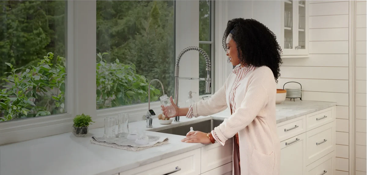 Una mujer con un cárdigan blanco y una blusa rosa llena un vaso con agua hidrogenada en el fregadero de la cocina. La cocina tiene armarios blancos y una gran ventana con vistas a una exuberante vegetación. Junto a ella hay varios vasos vacíos en una bandeja.