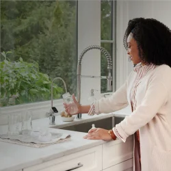 Una mujer con un cárdigan blanco y una blusa rosa llena un vaso con agua hidrogenada en el fregadero de la cocina. La cocina tiene armarios blancos y una gran ventana con vistas a una exuberante vegetación. Junto a ella hay varios vasos vacíos en una bandeja.