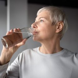 Una mujer mayor, de pelo corto y gris, que viste una camisa gris claro, bebe agua hidrogenada de un vaso transparente. Está de pie en una habitación con una iluminación suave y natural y un fondo neutro, mirando hacia su derecha.