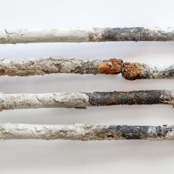 Primer plano de un elemento calefactor corroído y con abundantes incrustaciones sobre un fondo blanco. El metal está oxidado y cubierto de depósitos minerales blancos, lo que resalta el desgaste y el deterioro.