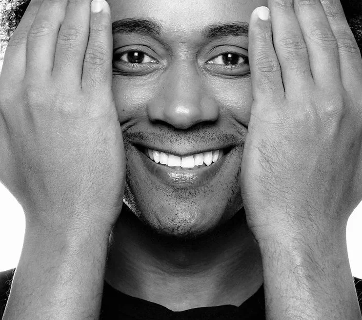 Imagen en blanco y negro de una persona sonriendo ampliamente, cubriéndose parcialmente el rostro con las manos, como si se refrescara con un chorro de agua de un purificador de agua. Los dedos están separados, capturando una expresión alegre.